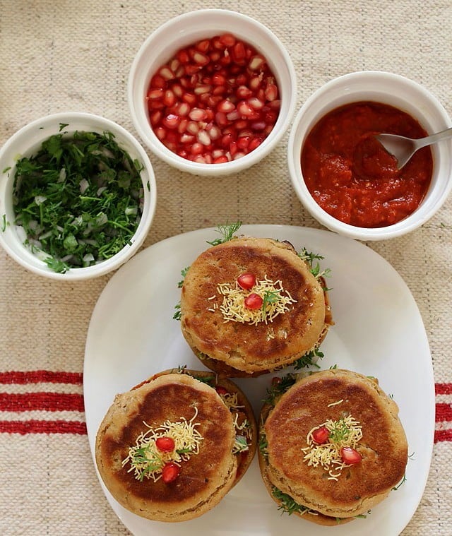 top shot of dabelis served on a square white plate with garnishes and red garlic chutney kept in bowls on the top. 