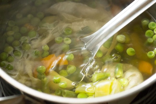 half cooked vegetables floating in water with a steel spoon inside