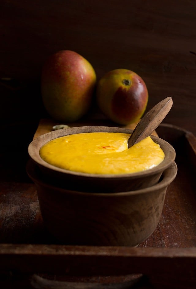 mango shrikhand served in a wooden bowl with wooden spoon inside with reddish yellow colored mangoes in the background