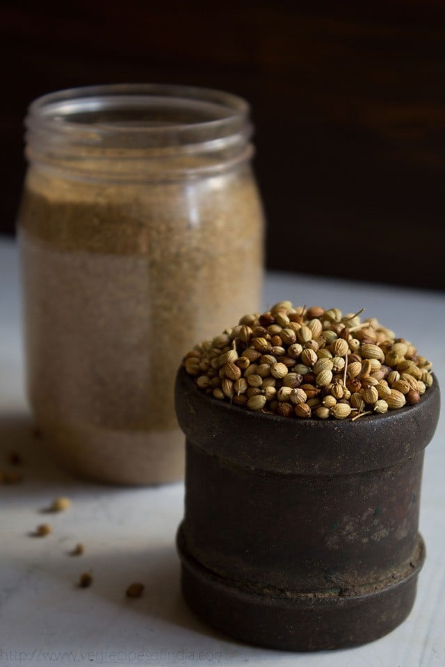 coriander seeds in a black iron measuring cup with a jar filled with ground coriander powder in the background