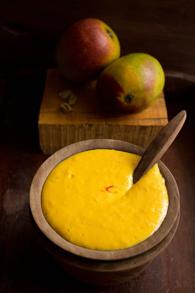 mango shrikhand served in a bowl with a wooden spoon inside and with fresh ripe mangoes in the background