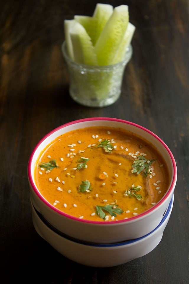 muhammara (smoky red pepper dip) in a white bowl with a red rim, garnished with a drizzle of olive oil, fresh coriander leaves and toasted sesame seeds.