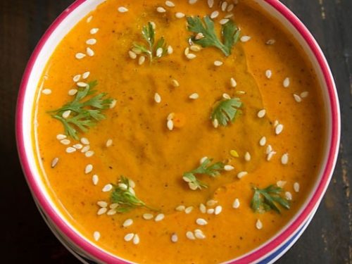 muhammara (roasted red pepper dip) in a white bowl with a red rim, garnished with a drizzle of olive oil, fresh coriander leaves and toasted sesame seeds.