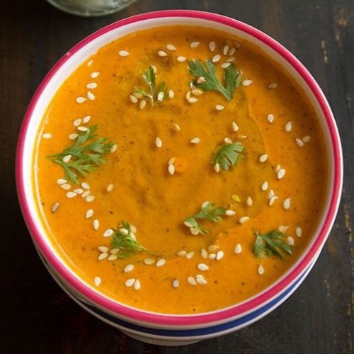 muhammara (roasted red pepper dip) in a white bowl with a red rim, garnished with a drizzle of olive oil, fresh coriander leaves and toasted sesame seeds.