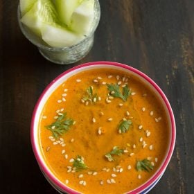 muhammara (red pepper dip) in a white bowl with a red rim, garnished with a drizzle of olive oil, fresh coriander leaves and toasted sesame seeds.