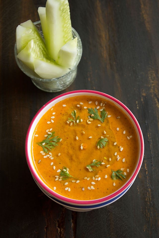 muhammara (red pepper dip) in a white bowl with a red rim, garnished with a drizzle of olive oil, fresh coriander leaves and toasted sesame seeds.