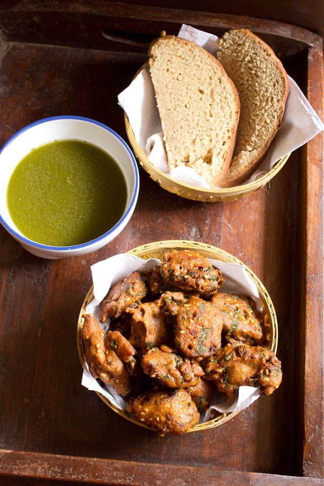 palak pakoda served in a bowl with a side of wheat bread slices and coriander chutney on a wooden board