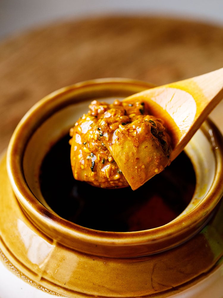 mango pickle scooped in a wooden spoon on top of a ceramic jar placed on a brown wooden board