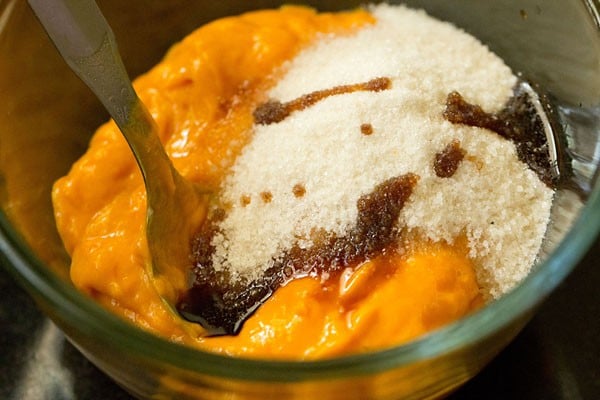 sugar and vanilla added to mango pulp in a glass bowl with a steel spoon in it