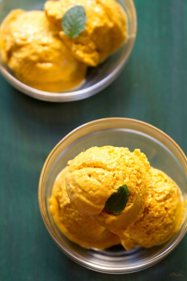 scoops of mango ice cream in two glass bowls garnished with a mint leaf on a green board