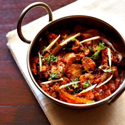 kadai mushroom garnished with coriander leaves, ginger juliennes and served in a steel kadai on a white napkin on a dark ebony colored board.