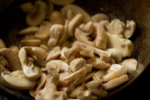 sliced white button mushrooms being sautéed in oil in the kadai