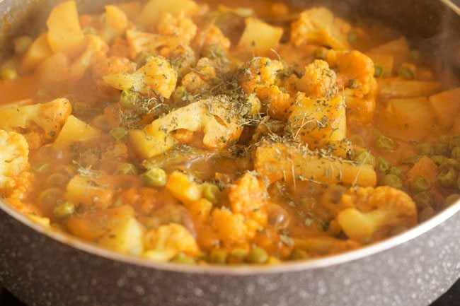 adding crushed kasuri methi to aloo gobi matar curry in pan