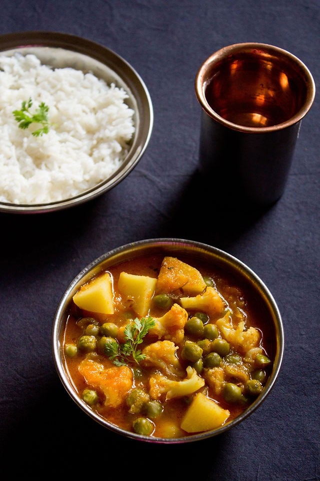 aloo gobi matar curry served in a steel bowl with a side of steamed rice in bowl and glass of water