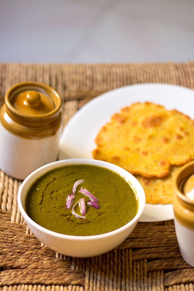 sarson ka saag in a white bowl topped with chopped onions and served with a piece of makki ki roti.