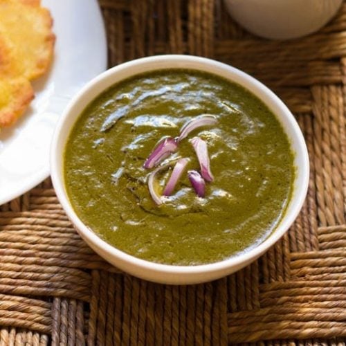 sarson ka saag in a white bowl on a rattan table with a white plate of makki ki roti to the side.