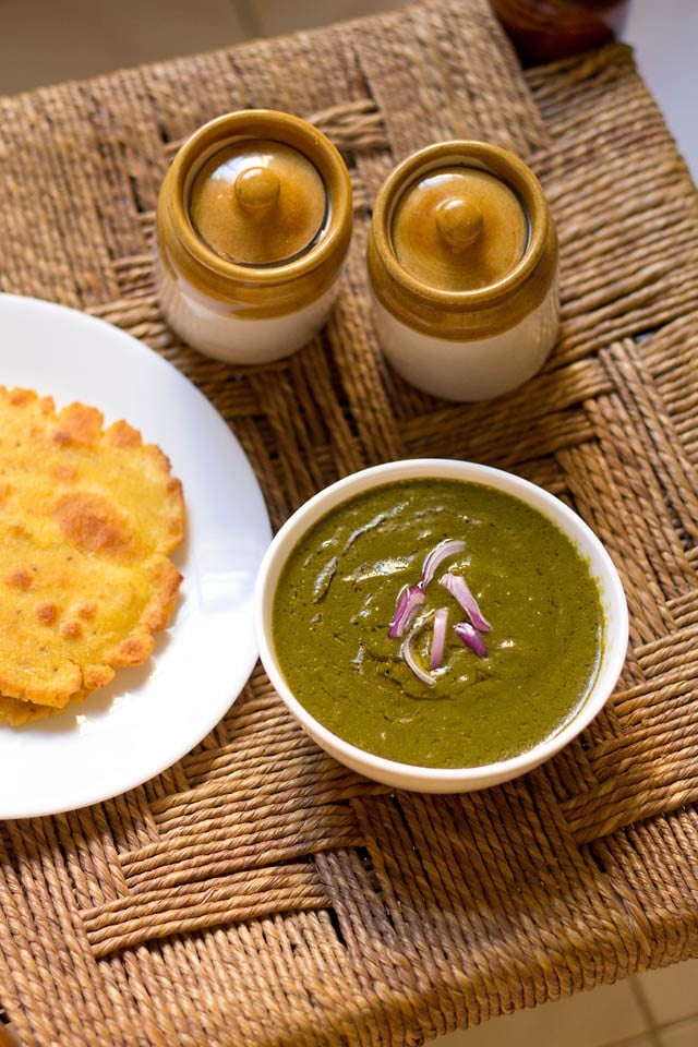 sarson ka saag in a white bowl on a rattan table with a white plate of makki ki roti to the side.