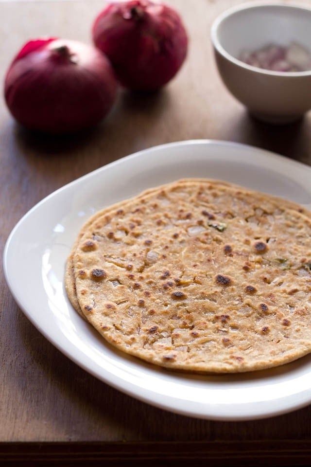 two onion paratha stacked in a square white plate with red onions and chopped onions, green chillies in a small white bowl in background