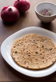 two onion paratha stacked on a square white plate with red onions and chopped onions, green chilies in a small white bowl in background.