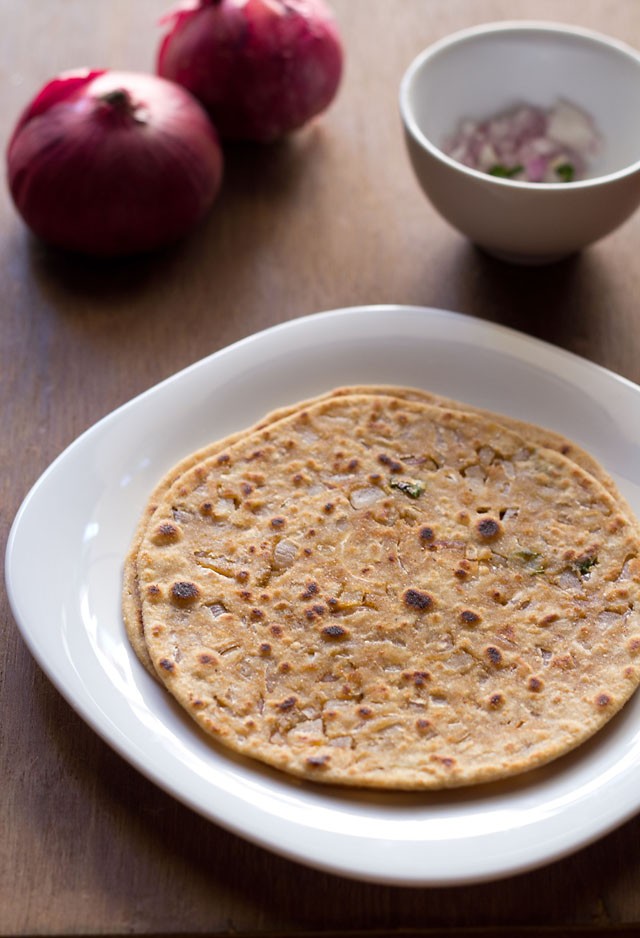 two onion paratha stacked in a square white plate with red onions and chopped onions, green chillies in a small white bowl in background