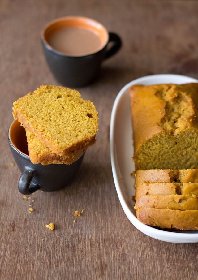 orange cake slices on an empty black cup with a half loaf of orange cake on a white oblong tray at left side and a black cup filled with tea on the top