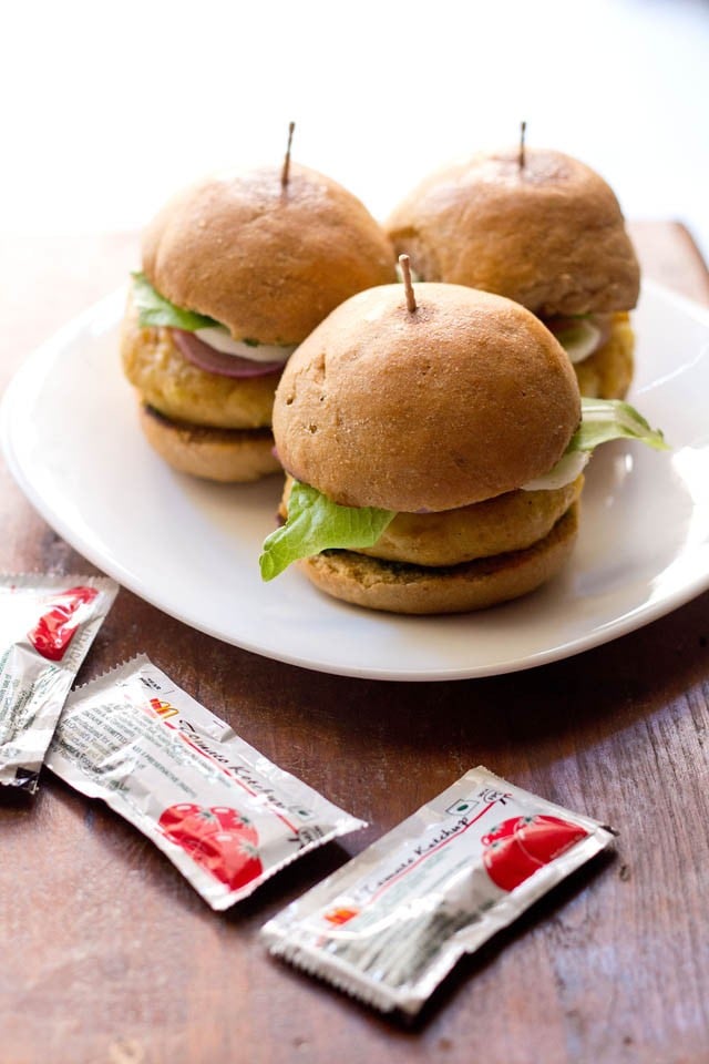 3 aloo tikki burgers on a white plate with packets of ketchup on the table.