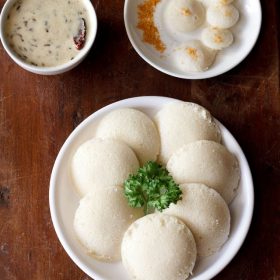 steamed idli with idli rava garnished with a coriander leaf and served on a white plate with a bowl of coconut chutney on the top left side and a plate of podi sprinkled idlis on the top right side.