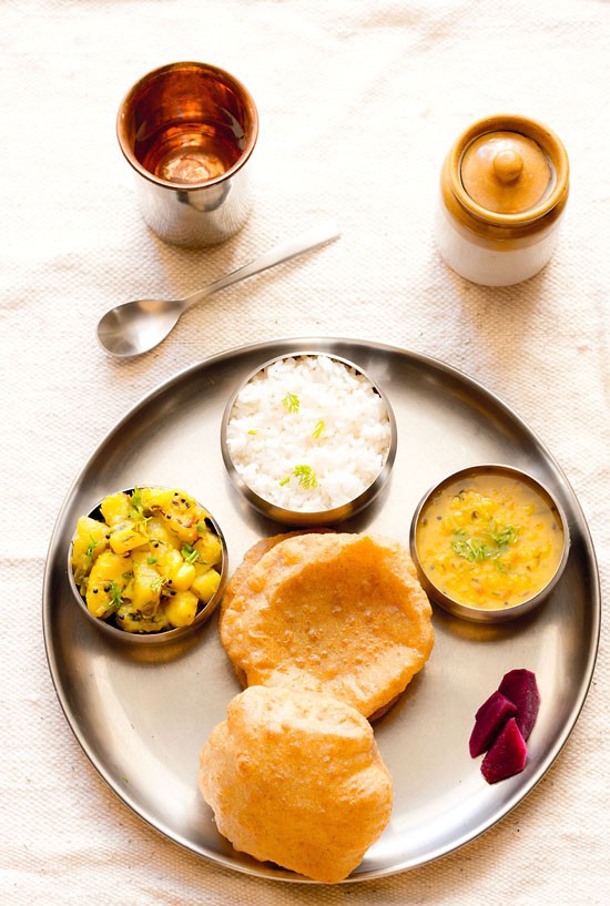 thali meal with bowls of steamed rice, moong dal, fresh puri, and batata bhaji.