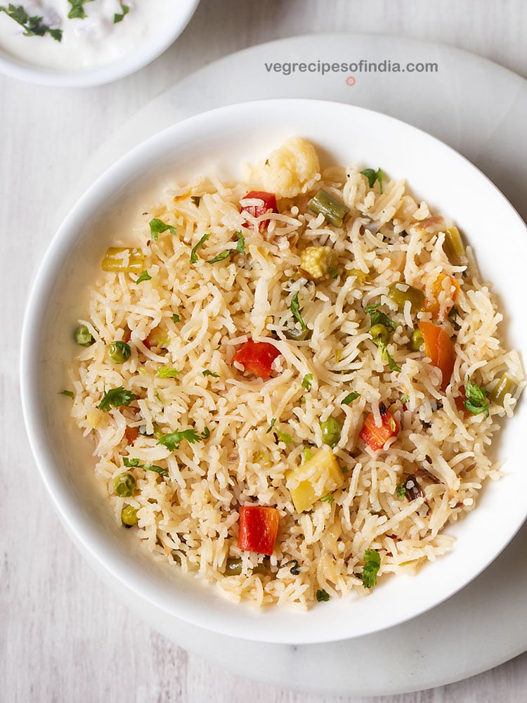 veg pulao served in a white shallow bowl with yogurt raita by the side