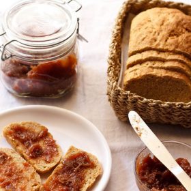 fig jam in a bowl with the jam spread on wheat bread in a white plate and a fig jam jar and loaf of bread in the background