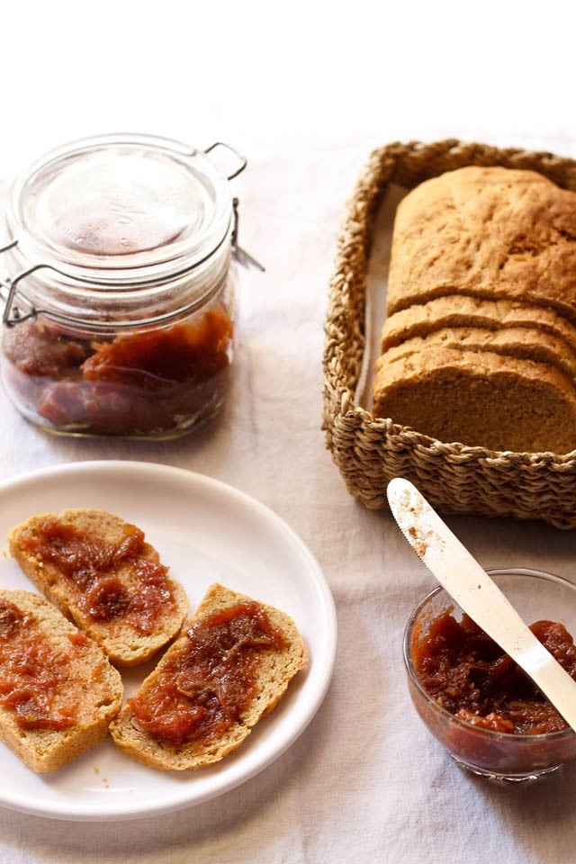 fig jam in a bowl with the jam spread on wheat bread in a white plate and a fig jam jar and loaf of bread in the background