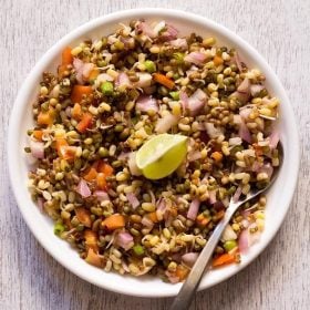 moong sprouts salad in a white plate with a lemon wedge in the center and a spoon on the side.