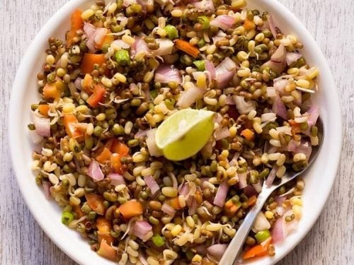 moong sprouts salad in a white plate with a lemon wedge in the center and a spoon on the side.