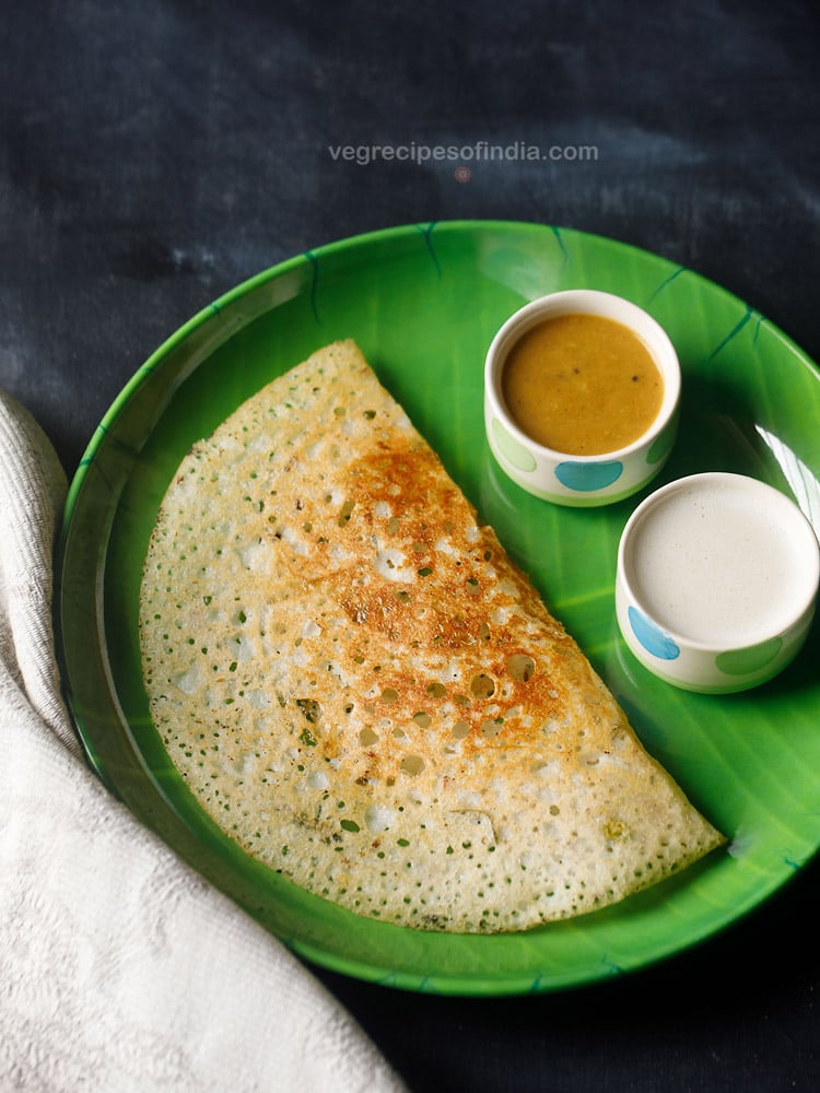 sooji dosa served in a green plate with a side of two bowls having sambar and coconut chutney on a dark black-blue board