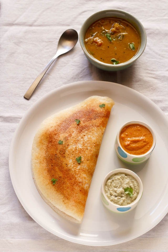 Mysore masala dosa is served on a plate with a small bowl of garlic chutney, a small bowl of coconut chutney, a bowl of sambar and a spoon placed in the upper left corner.  . 