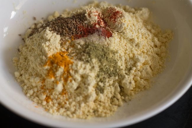 gram flour, turmeric powder, asafoetida, red chili powder, fennel powder and carom seeds added to a white mixing bowl. 