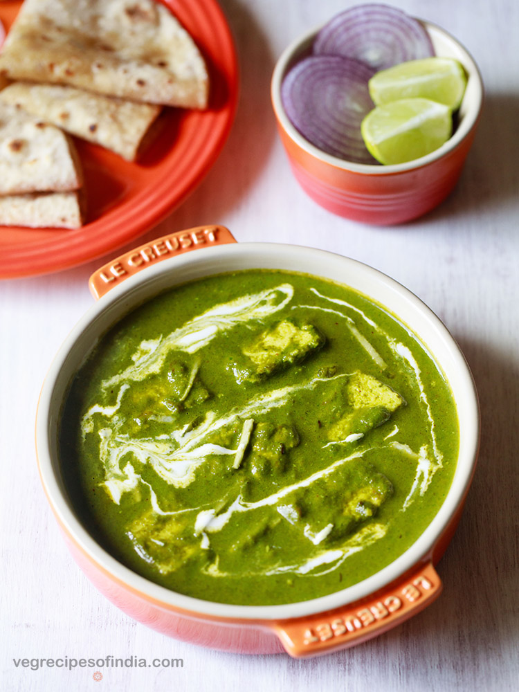 palak paneer topped with cream, garnished with ginger julienne and served in an orange bowl on a white board with roti kept on a dark orange plate on top left and a small orange bowl with round sliced onions and lemon wedges on top