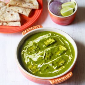 palak paneer topped with cream, garnished with ginger julienne and served in an orange bowl on a white board with roti kept on a dark orange plate on top left and a small orange bowl with round sliced onions and lemon wedges on top