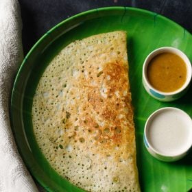 rava dosa geserveerd in een groen bord met aan de zijkant twee kommetjes met sambar en kokos chutney op een donker zwart-blauw bord