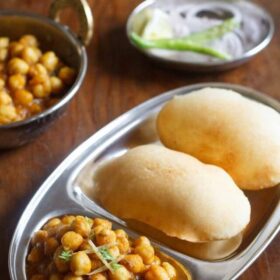 chole bhature served on a steel plate with a side of onion rings and green chilies, lemon in steel bowl and some extra chole in a copper bowl.
