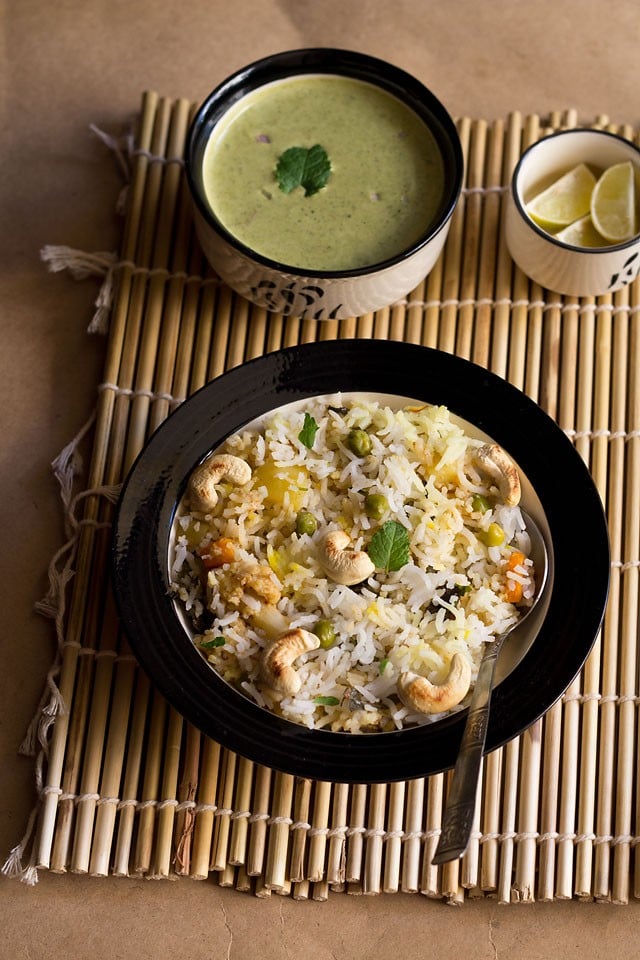 kashmiri biryani in a bowl on bamboo mat with raita and lemons in two bowls