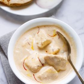 shahi paneer topped with some saffron strands served in a white bowl with a spoon inside the bowl on a white marble surface