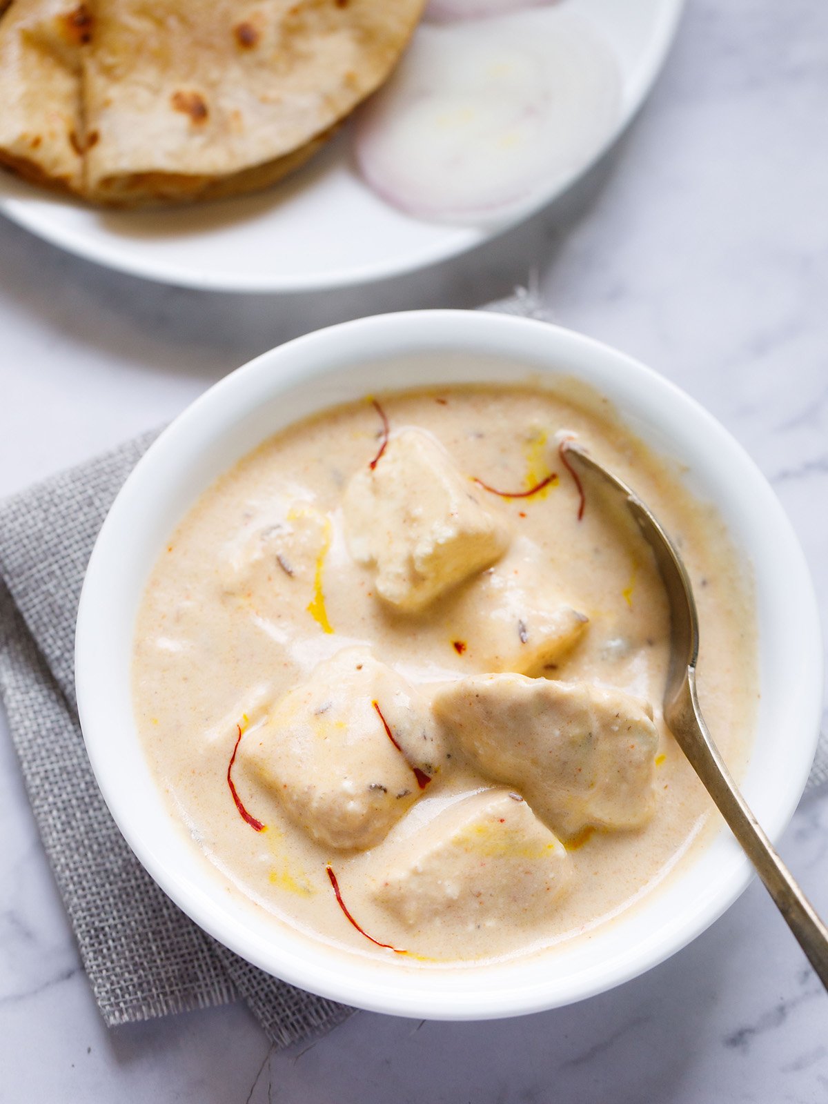 shahi paneer topped with some saffron strands served in a white bowl with a spoon inside the bowl on a white marble surface