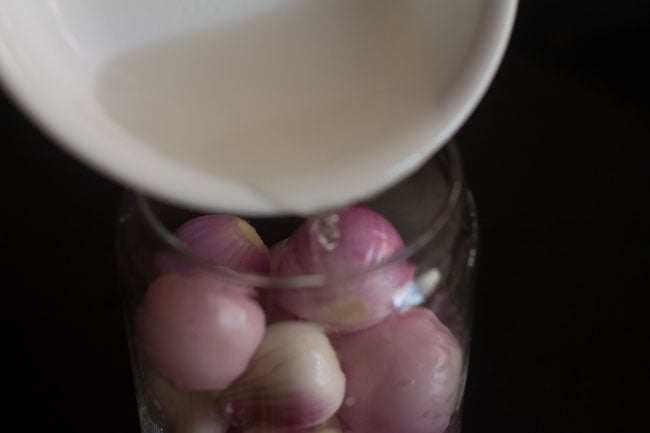 pouring vinegar-water mixture in the glass jar containing small onions 