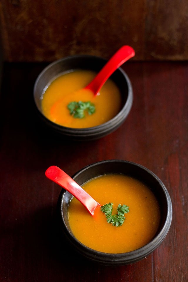 tomato carrot soup garnished with parsley and served in bowls with a red soup spoon.