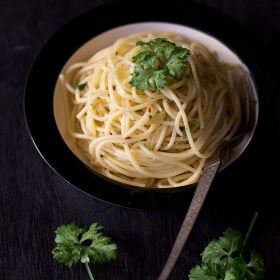 spaghetti aglio e olio garnished with parsley and served in a black rimmed white bowl with a fork kept on the right side.