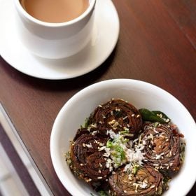 alu vadi garnished with grated coconut, coriander leaves and toasted sesame seeds in a white bowl with a cup of tea kept on the top left side.