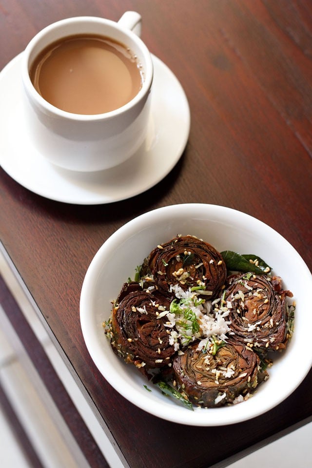 alu vadi garnished with grated coconut, coriander leaves and toasted sesame seeds in a white bowl with a cup of tea kept on the top left side.