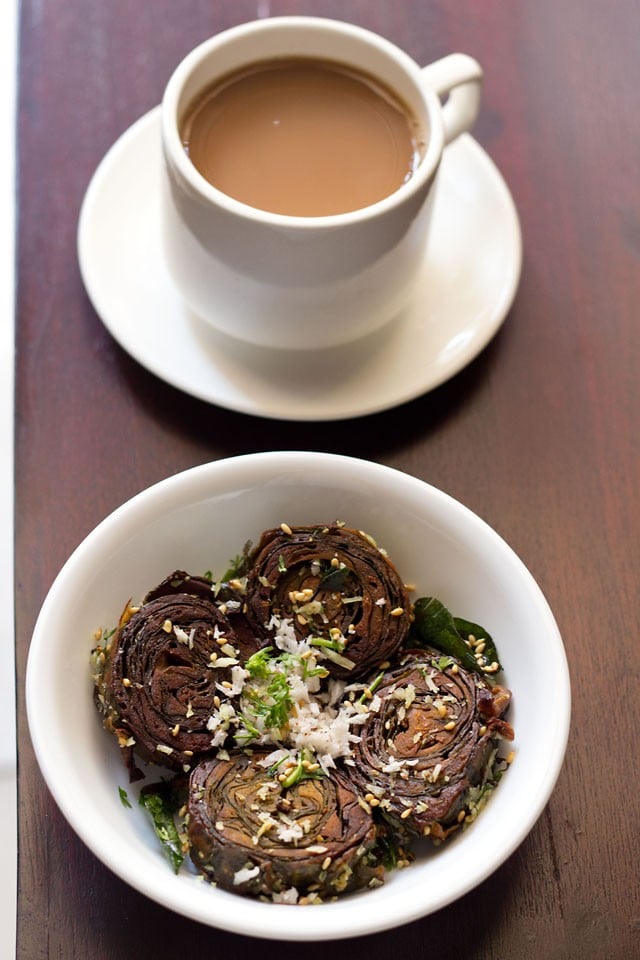 alu vadi garnished with grated coconut, coriander leaves and toasted sesame seeds in a white bowl with a cup of tea kept on the top side. 