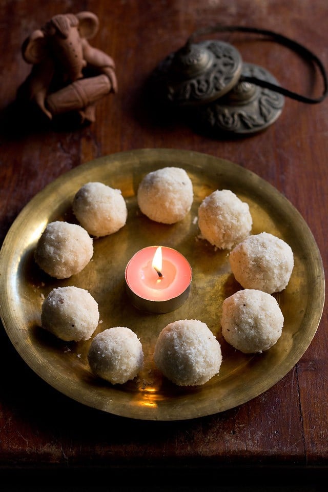 nariyal ke ladoo arranged in a circle with a pink tea light candle in the center on a brass plate with clay Ganesha and metal cymbals in the background